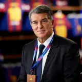 Prezes FC Barcelona, Joan Laporta, w garniturze, z identyfikatorem klubu na szyi. W tle widoczne koszulki piłkarskie.