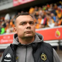 Skupiony trener Jagiellonii w kamizelce z herbem klubu, na tle trybun pełnych kibiców.