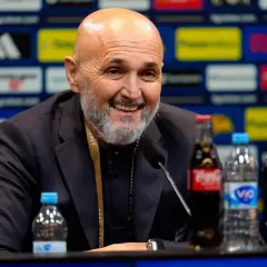 Uśmiechnięty trener Juventusu, z siwą brodą, siedzi przy stole z napojami.