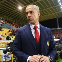 Sylvinho w granatowym garniturze i czerwonym krawacie, z emblematem na piersi, stoi na tle trybun stadionu.