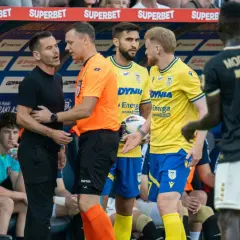 Piłkarze Arki Gdynia rozmawiają z trenerem. W tle widać ławkę rezerwowych, a kibice dyskutują o pogłoskach transferowych.