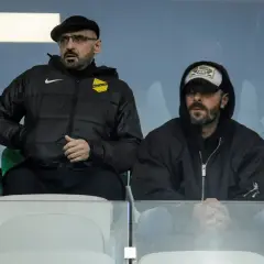 Dwóch mężczyzn siedzi na trybunach. Jeden z nich, w czapce i okularach, to właściciel wieczystej kraków.