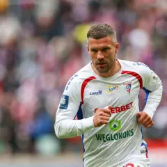 Piłkarz w białej koszulce z czerwonymi i niebieskimi detalami, biegnie po boisku. Mimo podolski wiek, widać jego determinację.