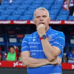 Trener Wisły Kraków w niebieskiej koszulce z białymi pasami, zamyślony, opiera dłoń na ustach.