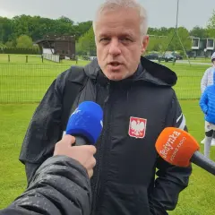 Jacek Jaroszewski, w czarnej kurtce z orłem, udziela wywiadu na boisku. W tle młodzi piłkarze.