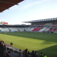 Widok na murawę i trybuny stadionu Cracovii. Na boisku trwa mecz, a trybuny zapełniają się kibicami.