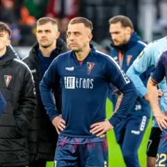 Piłkarze Pogoni Szczecin w ciemnych strojach, z widocznymi herbami klubu. W tle inni zawodnicy i sztab. Wszyscy wyglądają na zamyślonych, być może po trudnym meczu lub w oczekiwaniu na ważne pogoń szczecin - transfery.