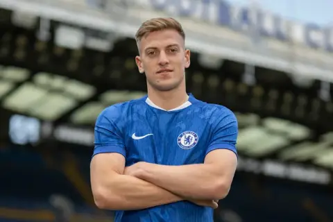 Młody piłkarz Chelsea w niebieskiej koszulce z herbem klubu, z rękami skrzyżowanymi na piersi.