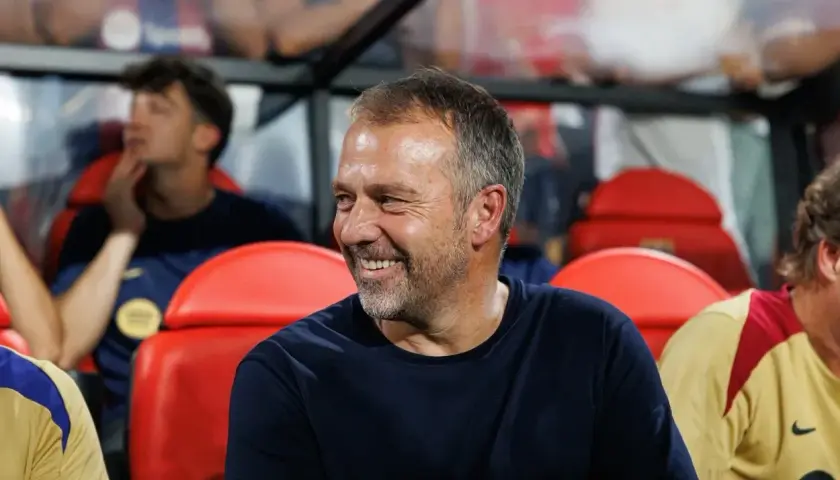 Uśmiechnięty mężczyzna, jeden z trenerów Barcelony, siedzi na ławce rezerwowych obok innych członków sztabu.