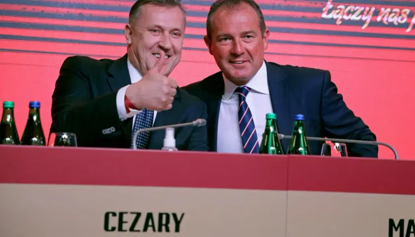 Cezary Kulesza, z synem, uśmiechnięci, z kciukiem w górze, podczas konferencji.