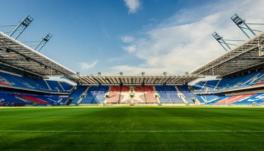 Pusta trybuna stadionu Wisły Kraków, z niebieskimi i czerwonymi siedzeniami tworzącymi wzór gwiazdy. Pojemność stadionu robi wrażenie.