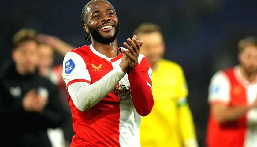 Uśmiechnięty Raheem Sterling w czerwono-białej koszulce, klaszczący. Jego statystyki w tym sezonie są imponujące.