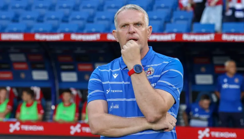 Trener Wisły Kraków w niebieskiej koszulce z białymi pasami, zamyślony, opiera dłoń na ustach.