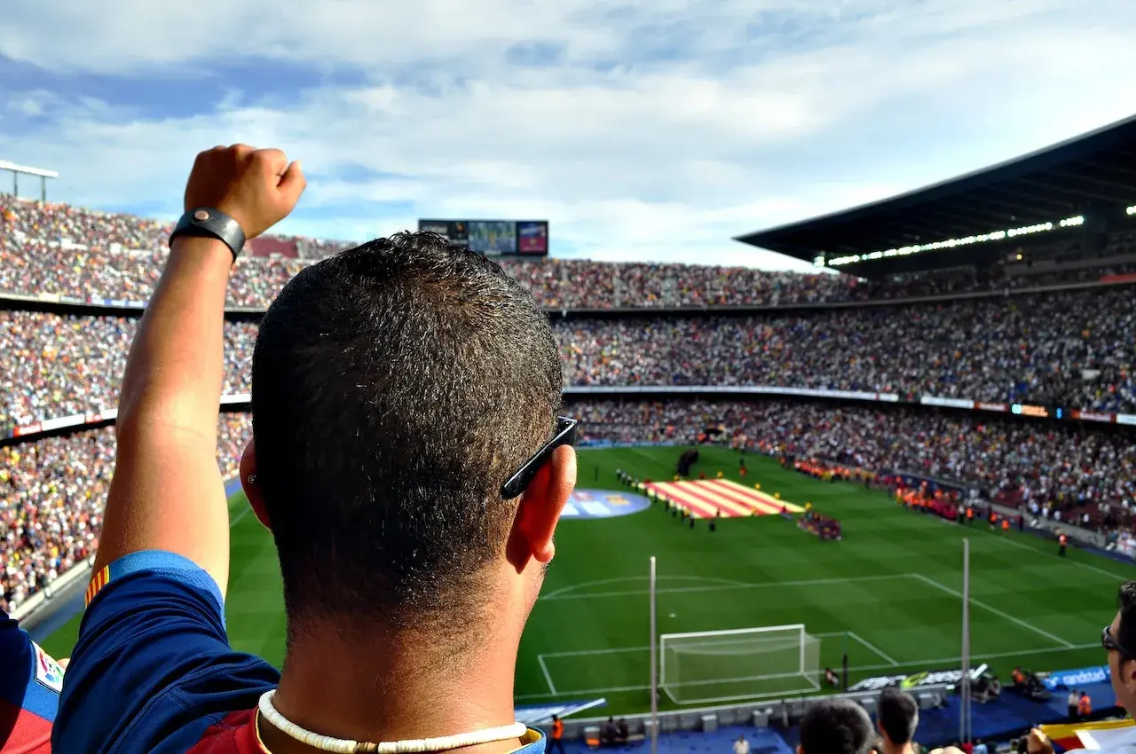 Zacięty kibic Barcelony z podniesioną pięścią na tle pełnego stadionu.