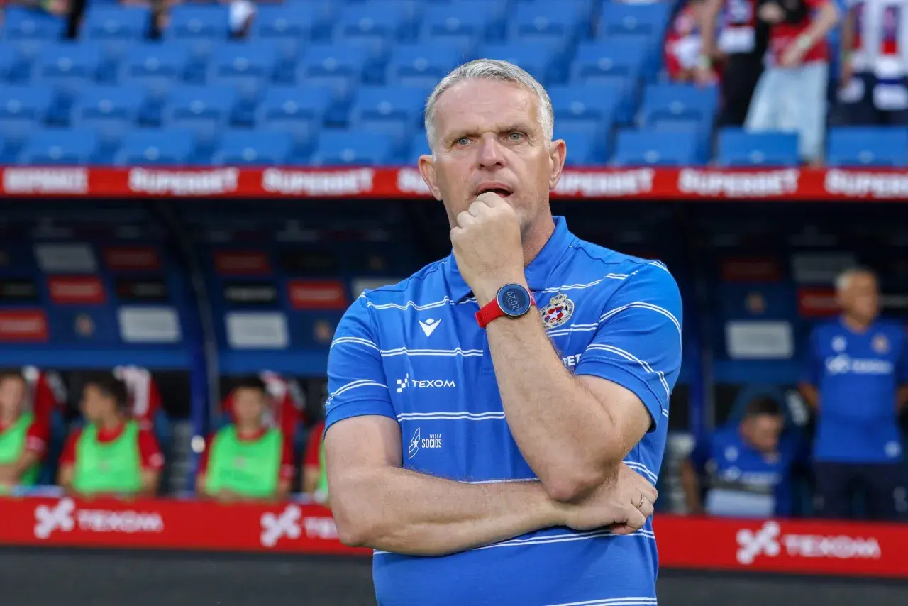 Trener Wisły Kraków w niebieskiej koszulce z białymi pasami, zamyślony, opiera dłoń na ustach.