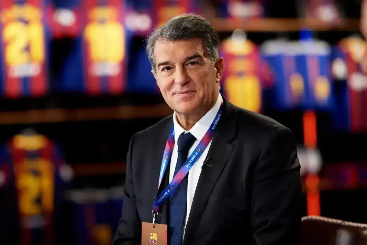 Prezes FC Barcelona, Joan Laporta, w garniturze, z identyfikatorem klubu na szyi. W tle widoczne koszulki piłkarskie.
