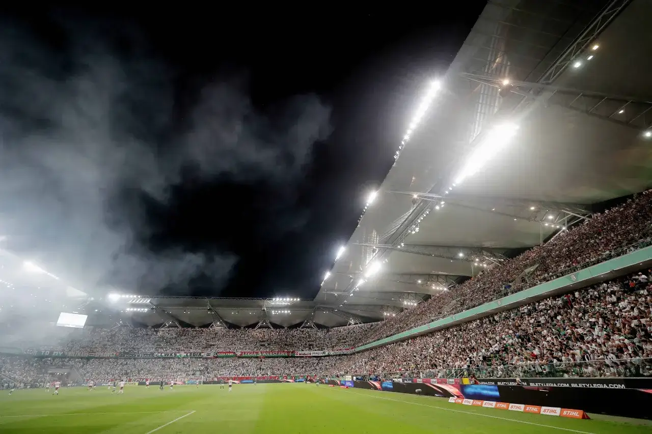 Stadion Legii Warszawa, pełen kibiców w białych koszulkach, z dymem unoszącym się w powietrzu. To obraz jednego z warszawskich klubów piłkarskich.