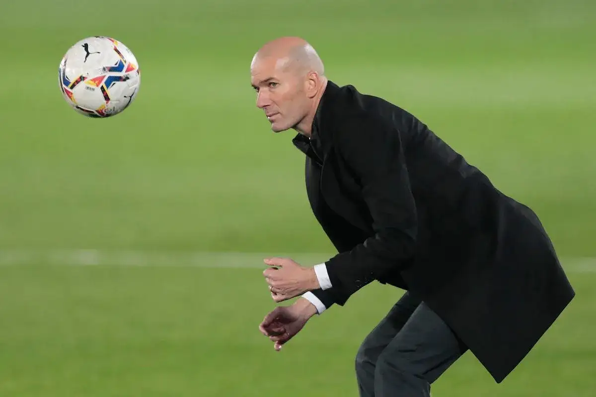 Zinedine Zidane, w czarnym płaszczu, pochyla się, obserwując lecącą piłkę. Jego statystyki jako trenera budzą podziw.