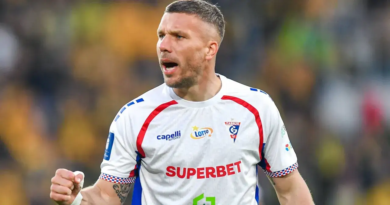 Piłkarz w białej koszulce z czerwonymi i niebieskimi detalami, zaciśnięta pięść, widać tatuaż. To podolski wiek, ale wciąż w formie.