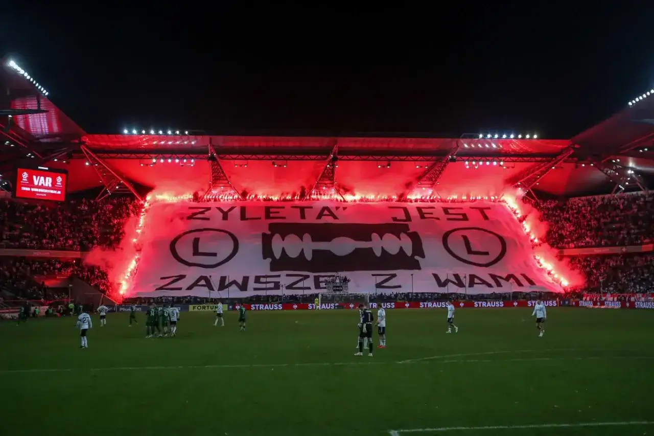 Trybuny stadionu Legii rozświetlone czerwonymi racami, na środku ogromny transparent z żyletą i napisem 