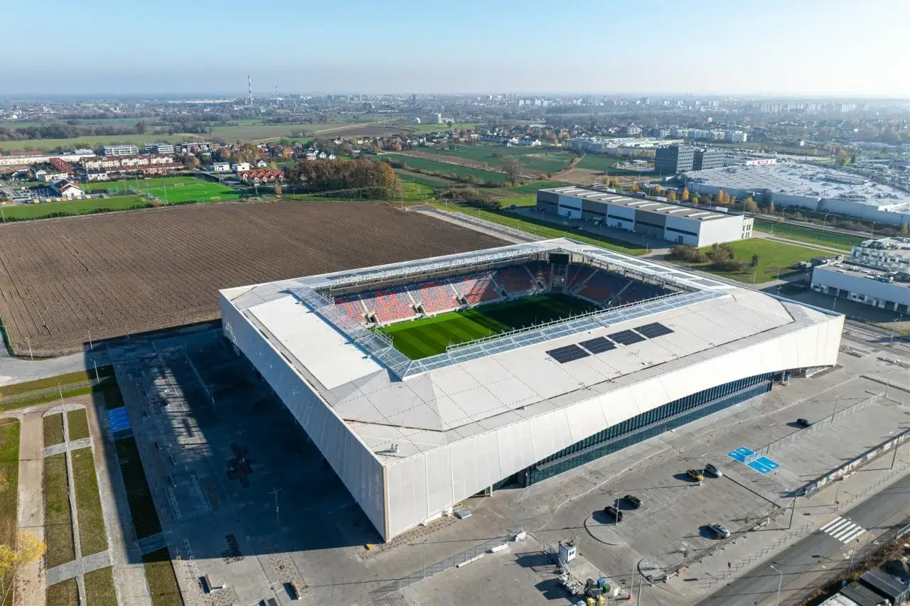 Nowoczesny stadion Odra Opole z zieloną murawą i pomarańczowo-niebieskimi trybunami, otoczony polami i budynkami przemysłowymi.