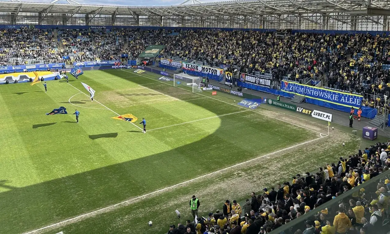 Stadion Lublin wypełniony kibicami w żółto-czarnych barwach. Na murawie osoby niosące flagi, przygotowujące się do meczu.