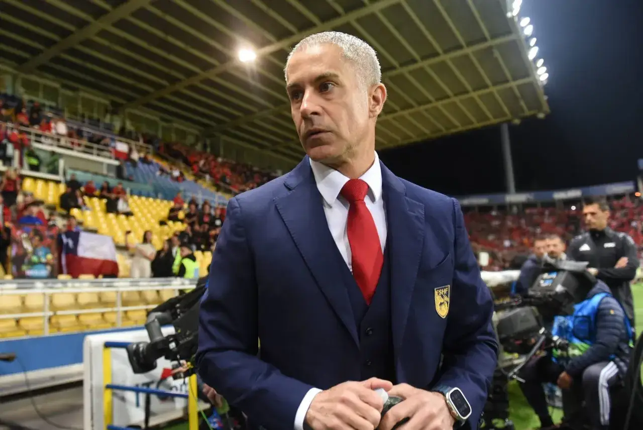 Sylvinho, w granatowym garniturze i czerwonym krawacie, stoi na tle trybun stadionu, gdzie kibice wznoszą flagę Chile.