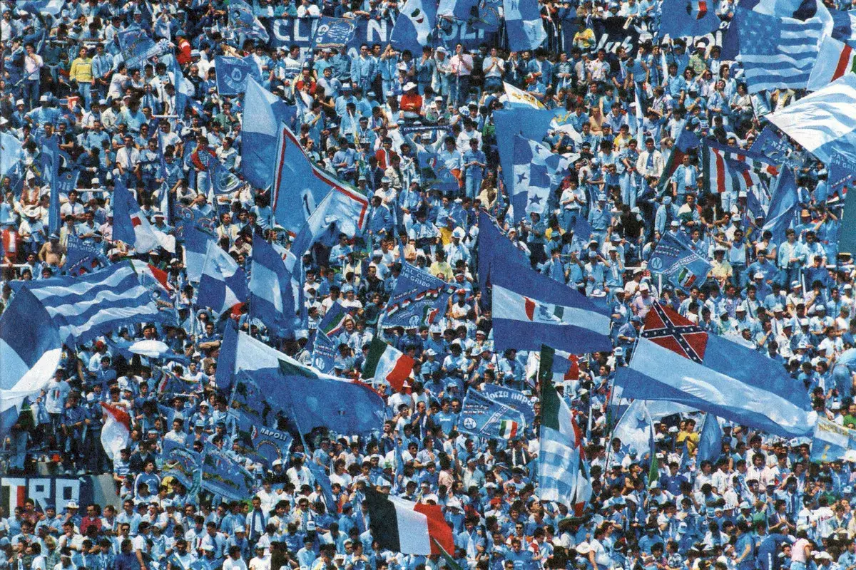 Tłum kibiców SSC Napoli, włoskiego klubu piłkarskiego, świętuje na stadionie, machając licznymi niebieskimi flagami.
