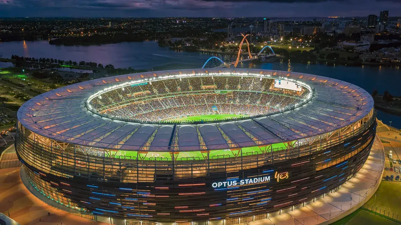 Optus Stadium, uznawany za najładniejszy stadion na świecie, lśni nocą w blasku świateł, otoczony wodą i miastem.